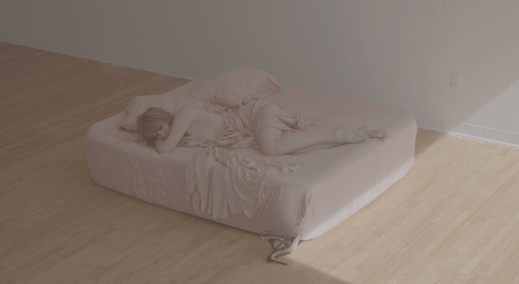 A muted image shows Jordan King gazing at the camera as she lays sprawled across an unmade bed entangled in a dusty rose sheet.